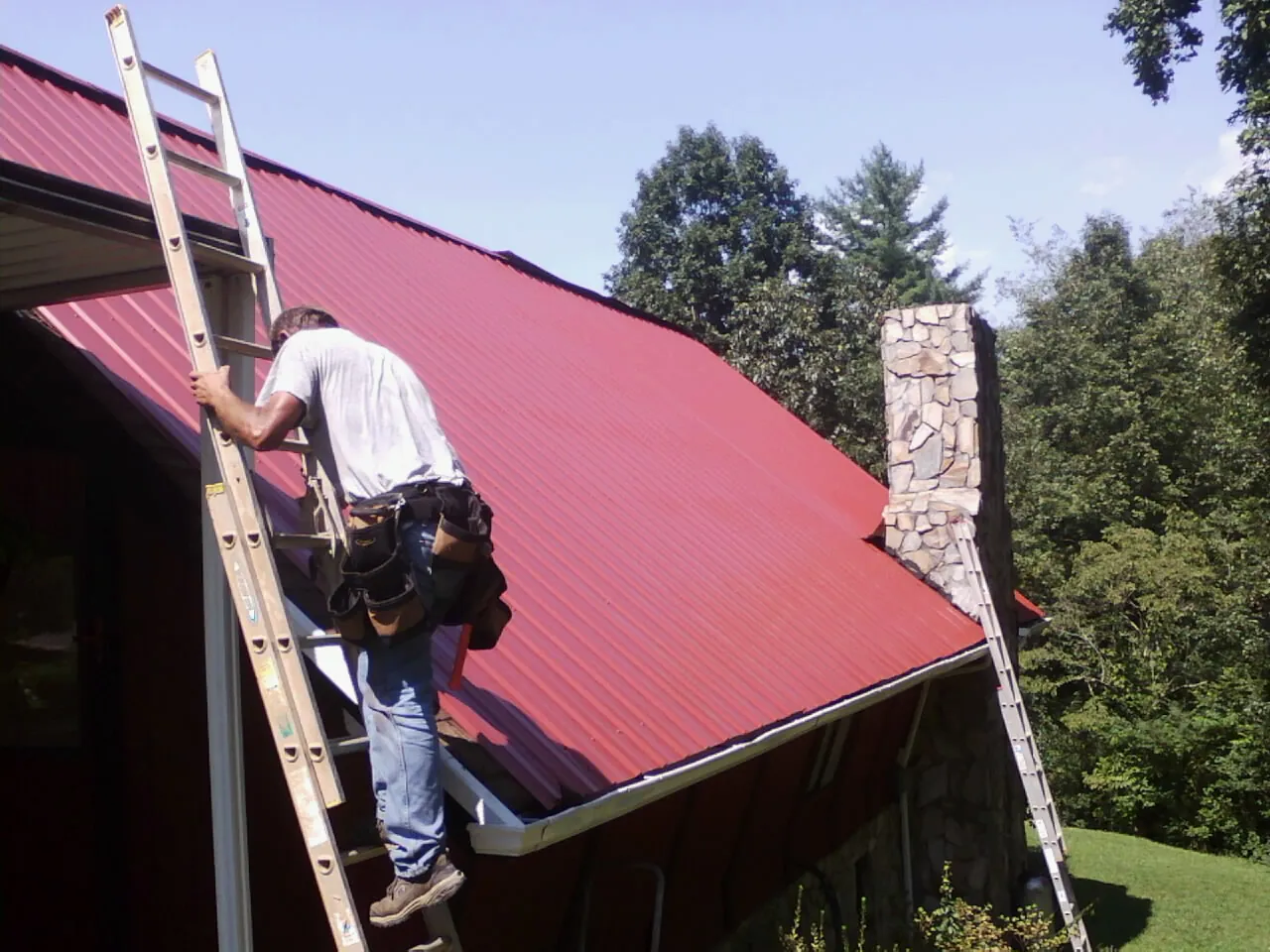 Roofer on a ladder beside a red metal roof and stone chimney — Soffit & Fascia Repair by Roofing Brothers in Center Point